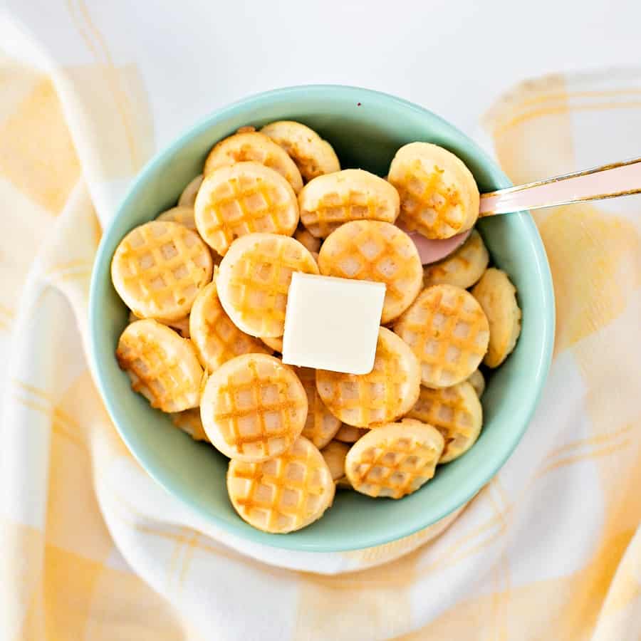 mini waffles in a blue green bowl with a pat of butter on top with a pink spoon inside with a yellow tea towel around the bowl