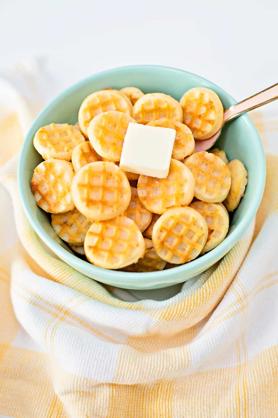 mini waffles in a blue green bowl with a pat of butter on top with a pink spoon inside