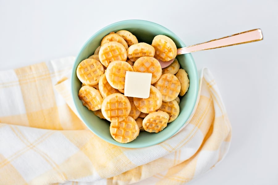 mini waffles in a blue green bowl with a pat of butter on top with a pink spoon inside with a yellow tea towel around bowl