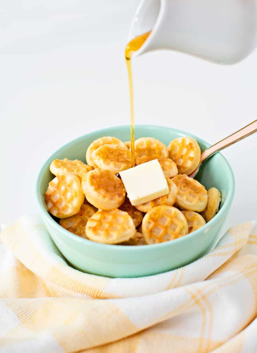 syrup being poured over mini waffle pancake cereal in a bowl